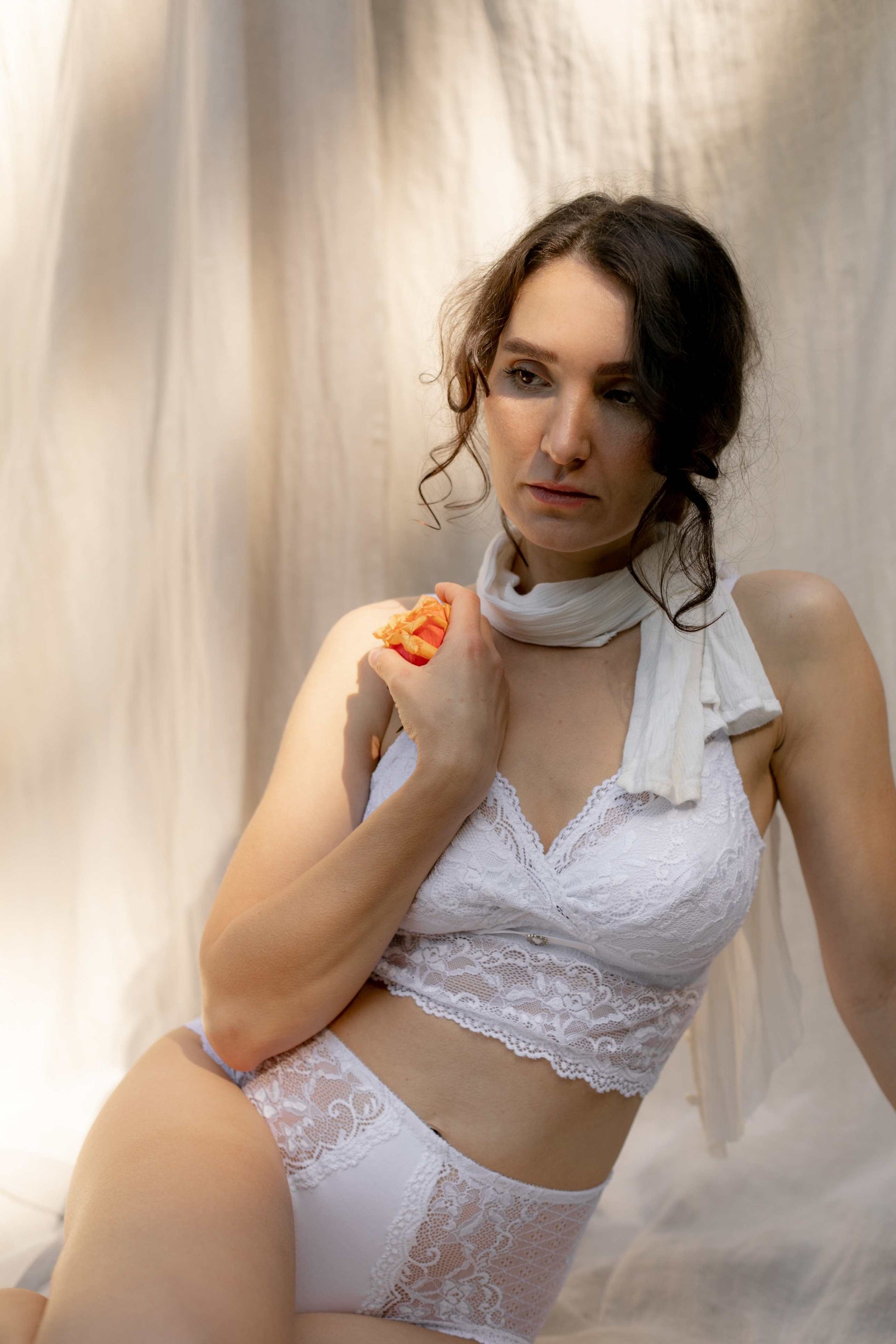 woman sitting  posing with orange flower in hand wearing white lace long line bra and white high rise lace panty